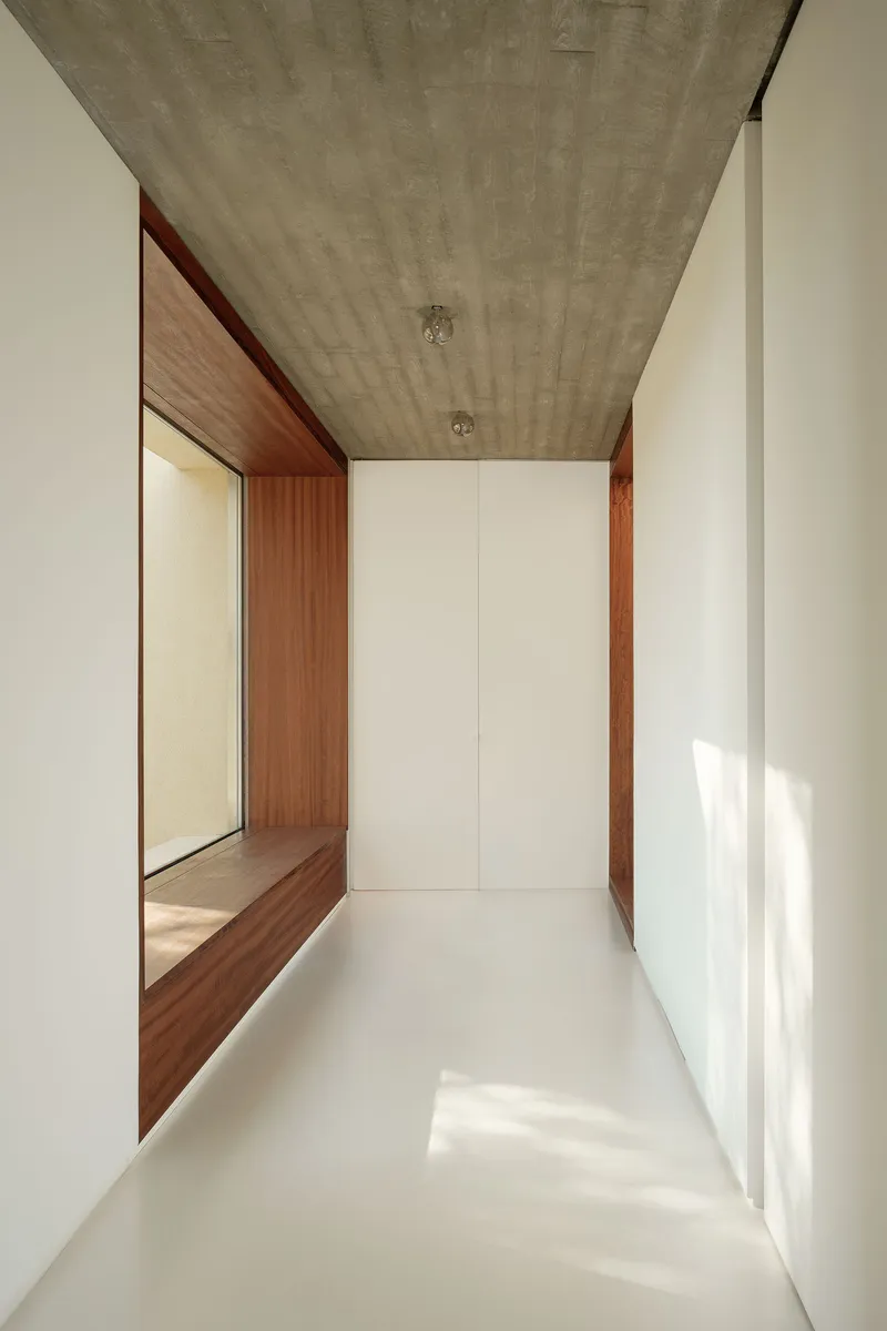 Corridor with exposed concrete ceiling, white walls, and timber-framed opening receiving afternoon sunlight