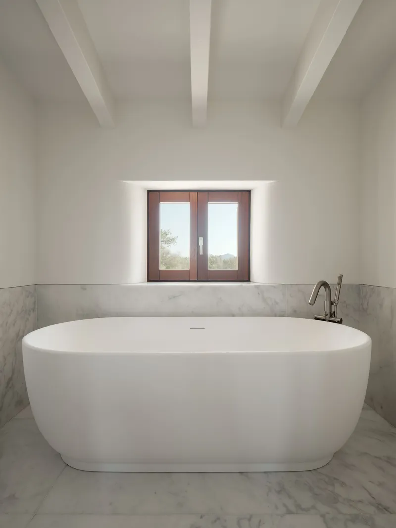 Freestanding oval bathtub beneath a deep-set timber-framed window with exposed ceiling beams