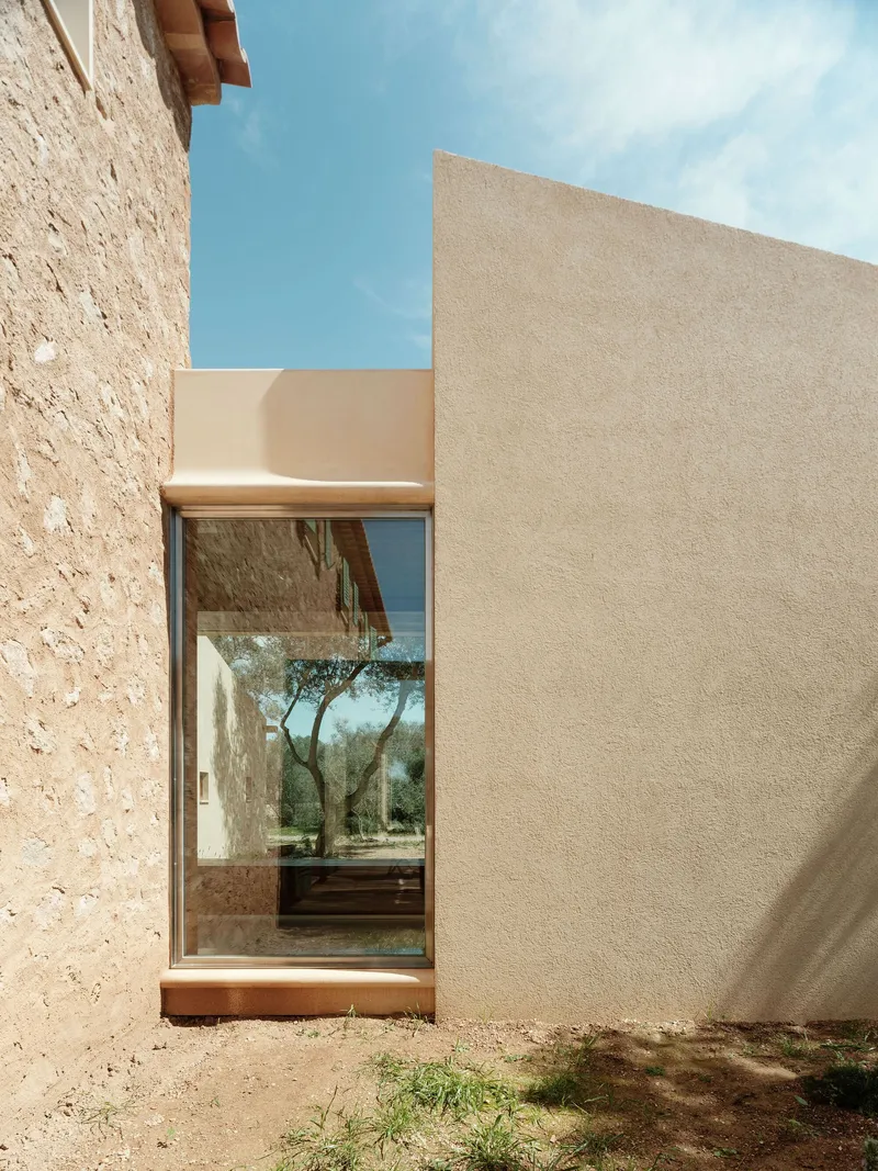 Glazed passage between stone and smooth plaster walls framing views of the olive grove beyond