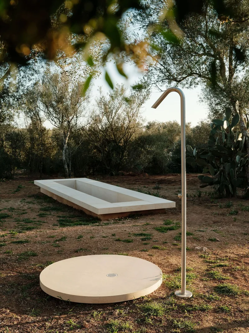 Outdoor shower with circular stone platform and curved metal fixture beside rectangular pool among olive trees