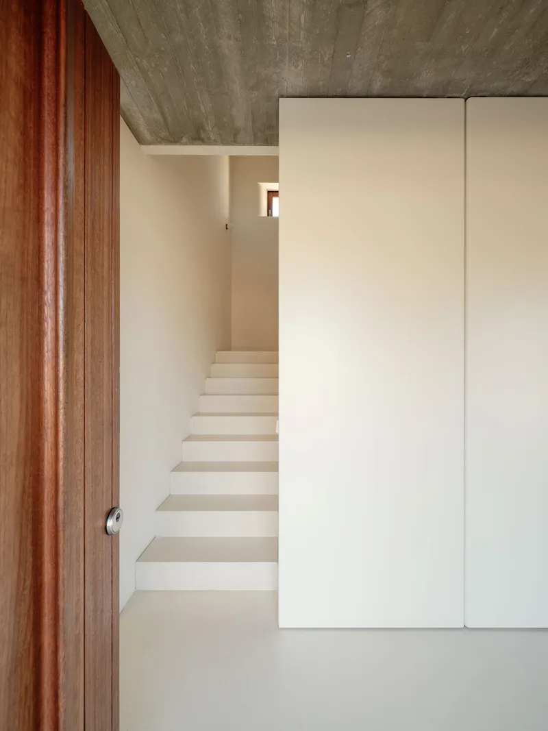 Plastered staircase rising between white walls beneath board-formed concrete ceiling