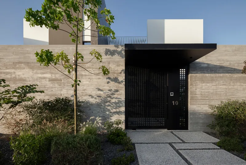 Black perforated metal entrance door with canopy overhang framed by planted beds and a young tree