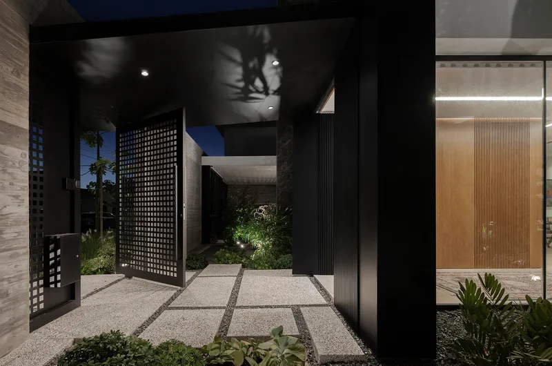 Entry courtyard at dusk with lattice pivot door, black walls and illuminated timber feature door beyond