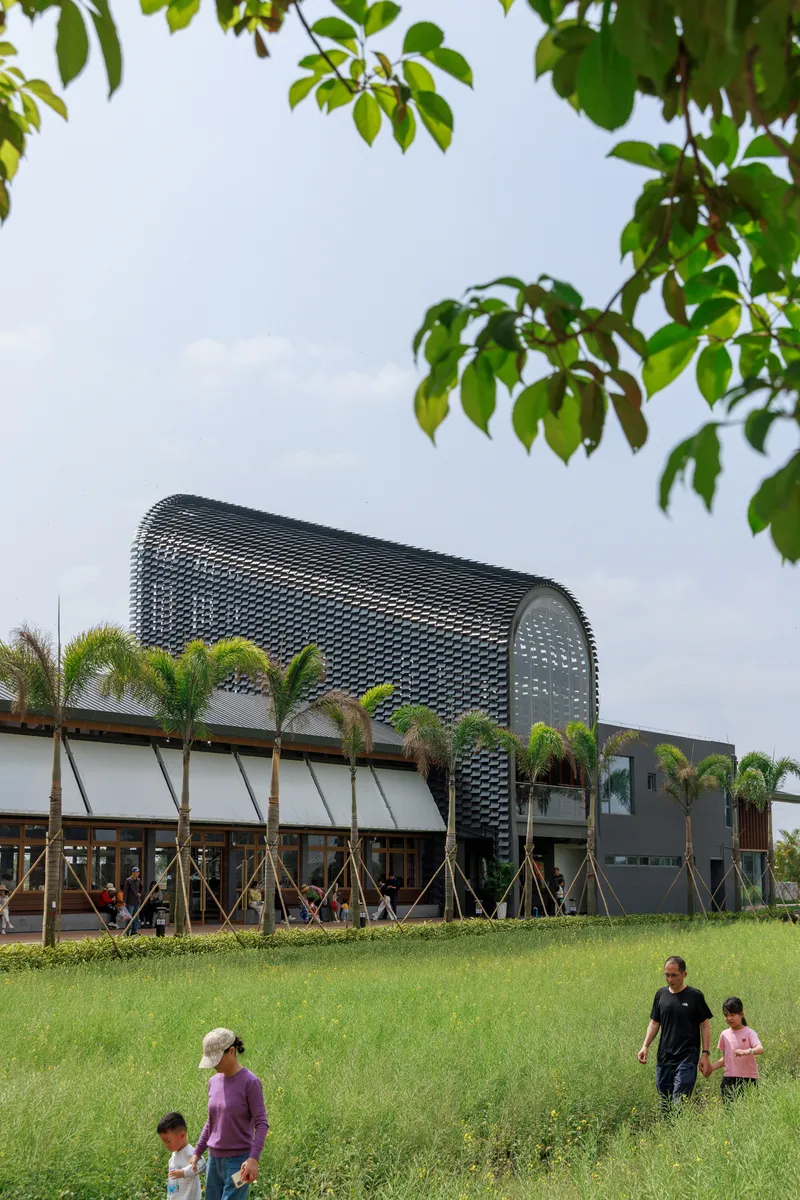 Perforated metal barrel vault rising above horizontal wing with palm trees and families on grassy lawn