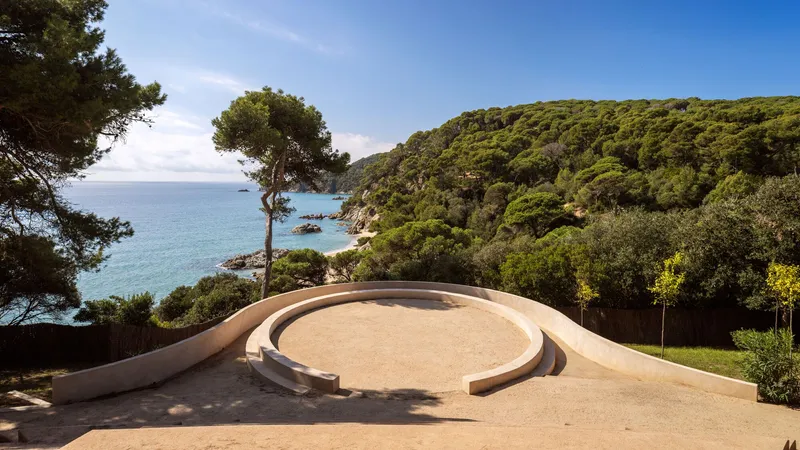 Curved amphitheatre with sand-colored terraces overlooking a forested coastal hillside and the sea