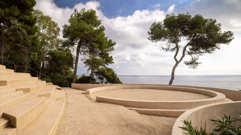 Circular sunken courtyard with stepped sand-colored walls framed by pine trees and cloudy sky