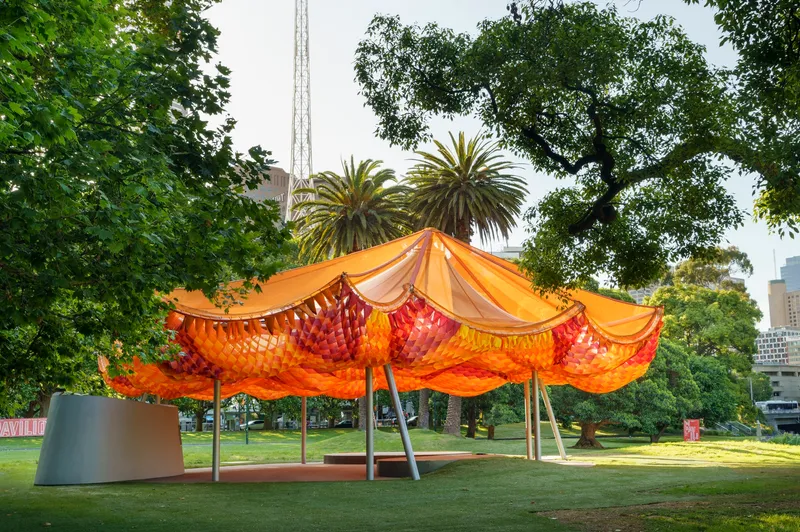 Tensile canopy with orange woven panels supported by angled columns in a park with palm trees