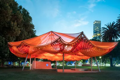 The image showcases a contemporary pavilion in an urban park setting. The structure features a strik