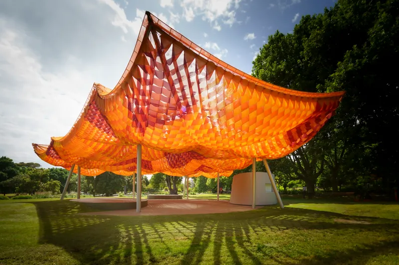 Side view of the scalloped canopy with woven panels creating dappled shadows on the paved ground