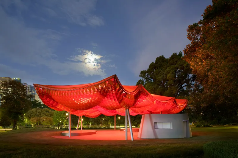 Pavilion with undulating red canopy on white columns in a park at twilight