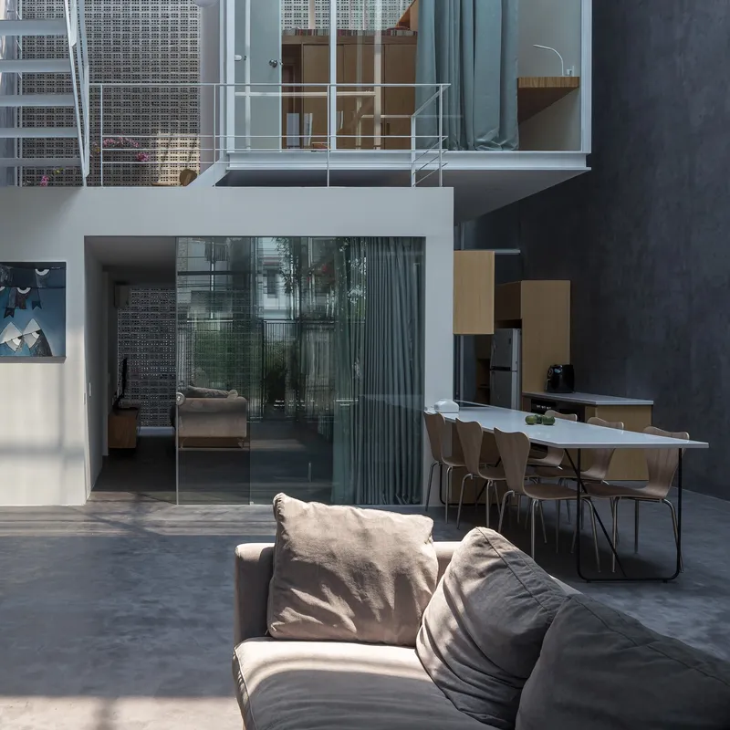 Double-height interior courtyard with glazed sliding doors, white steel balcony railing, and sunlight casting sharp shadows on polished concrete floor