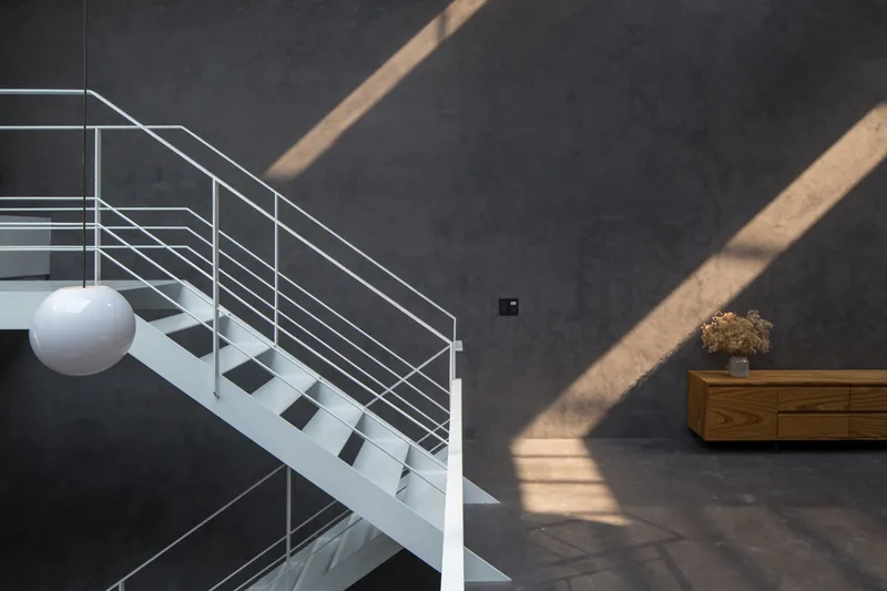White steel staircase with horizontal cable railings against dark grey wall, with angular sunlight patterns crossing the surface