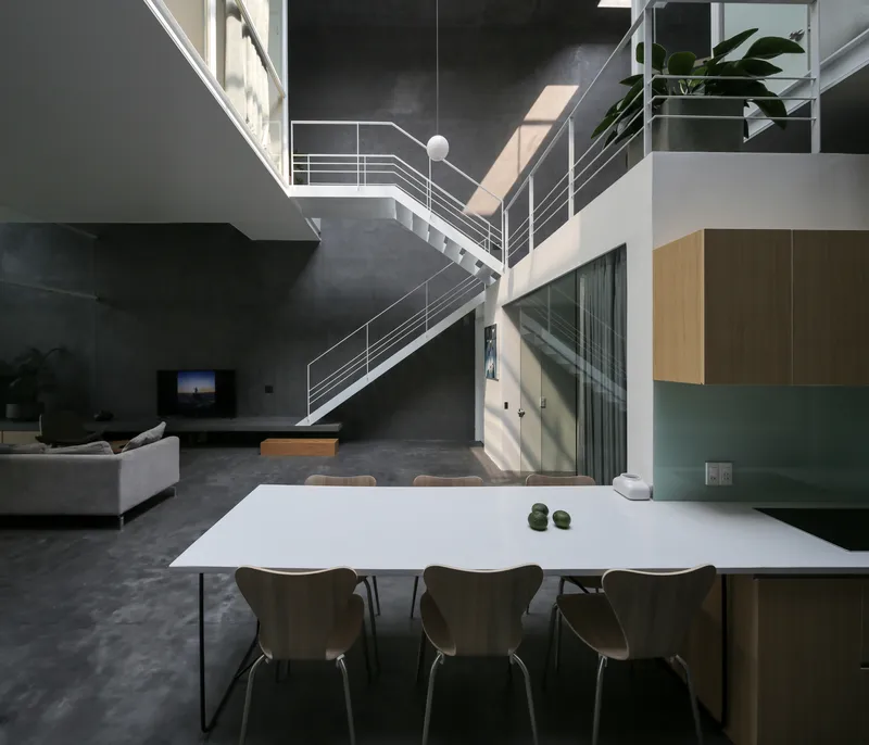 Open-plan dining area with white table beneath steel staircase and double-height concrete walls