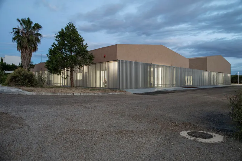 Corner view of the corrugated metal base and stucco upper volume with palm trees at dusk