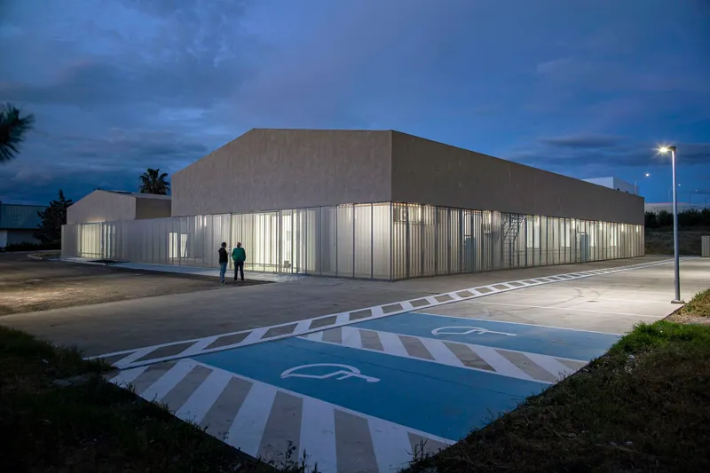 Illuminated translucent corrugated facade wrapping the building corner with two figures standing outside at twilight