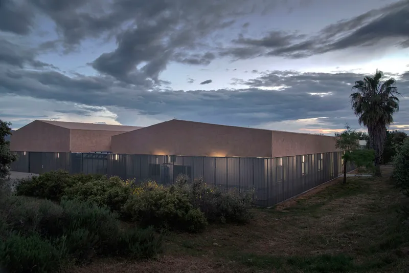 Backlit corrugated metal cladding beneath tan stucco volumes with native planting and stormy evening skies