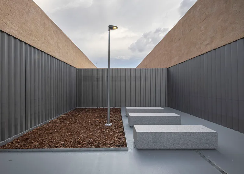 Enclosed courtyard with planted bed, concrete benches, and street lamp framed by ribbed metal walls