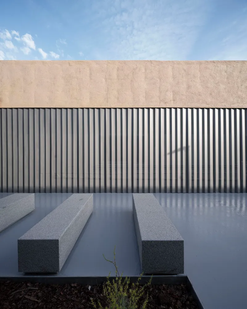 Courtyard with grey concrete benches and vertical metal siding below a beige plaster parapet under blue sky