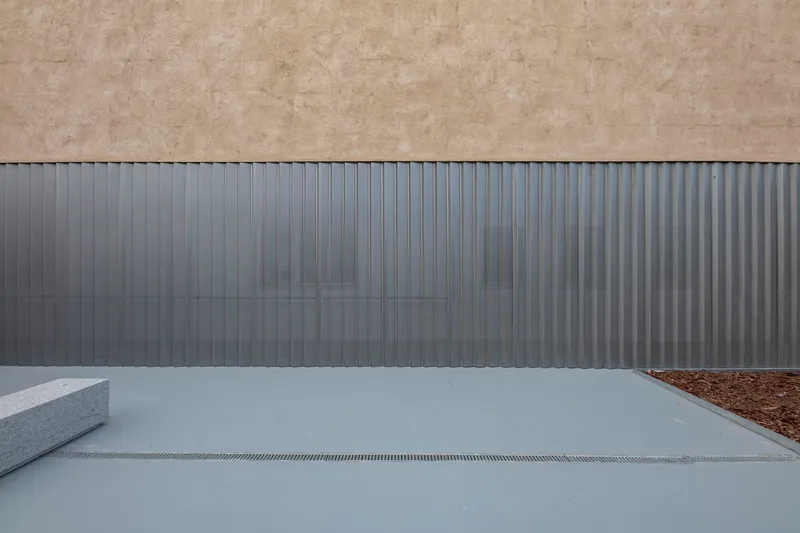 Close-up of vertical corrugated metal facade meeting concrete paving with linear drainage channel and planted bed