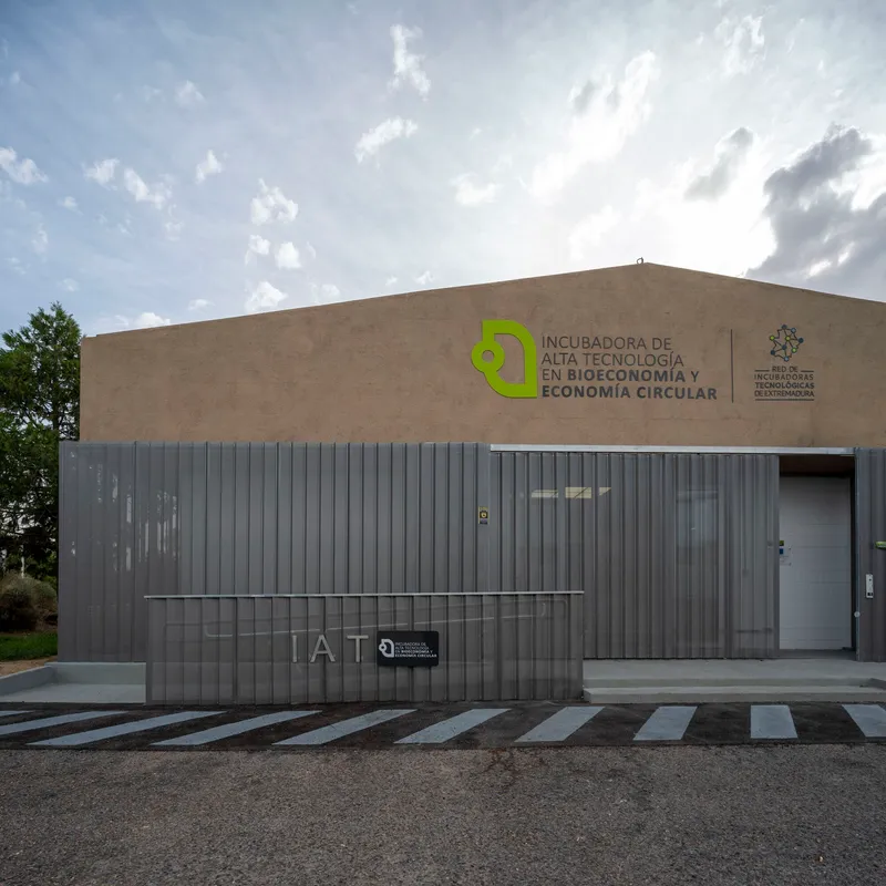 Street facade combining tan stucco upper volume with vertical corrugated metal cladding and signage under midday clouds