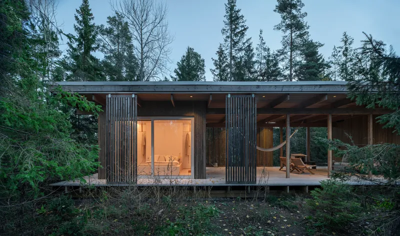 Flat-roofed timber cabin with vertical slatted screens and glazed openings nestled among evergreen trees at dusk