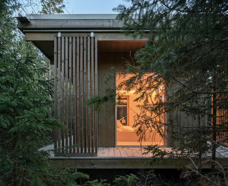 Corner module with vertical timber screen and glazed walls illuminated at dusk among conifers