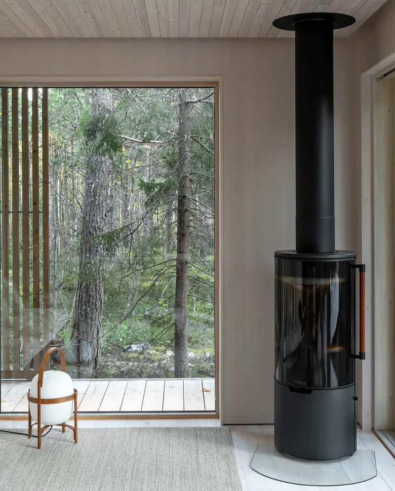 Freestanding black cylindrical wood stove beside a full-height window overlooking the forest