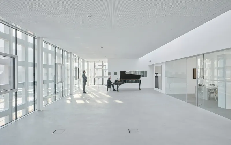 Open white interior with floor-to-ceiling glazing, a grand piano and two people in soft daylight
