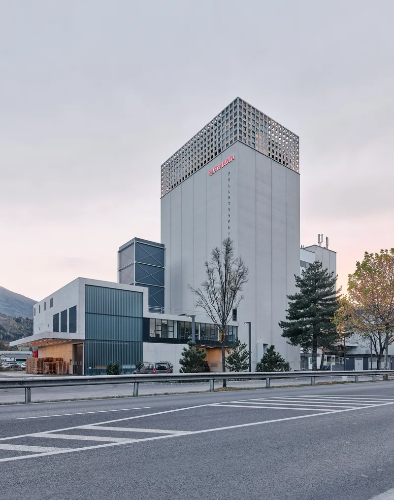 Oblique view of the concrete tower with latticed top rising above low-rise industrial volumes at dusk