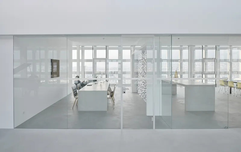 View through frameless glass partitions into the white workspace with pendant light and distant windows