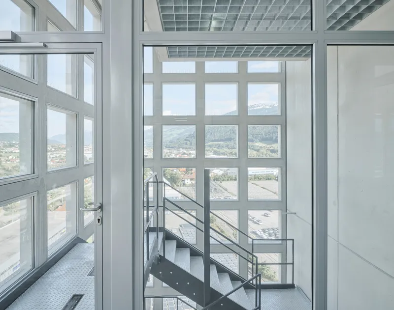 Steel staircase with wire mesh railing rising through a white-framed glass curtain wall overlooking distant mountains