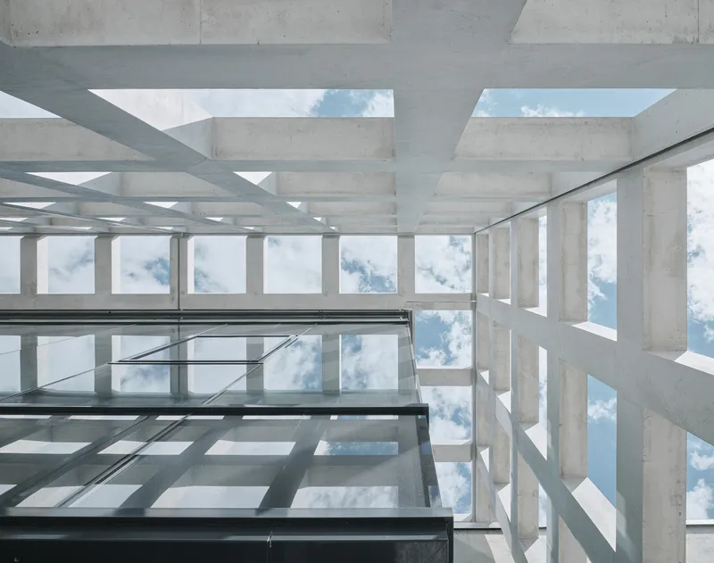 Upward view through a concrete waffle slab ceiling with glass floor panels revealing sky and clouds