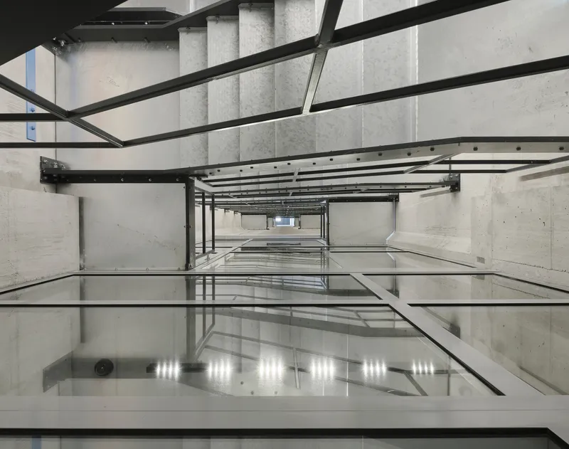 Looking down through a concrete elevator shaft with steel beams and reflective glass floor panels
