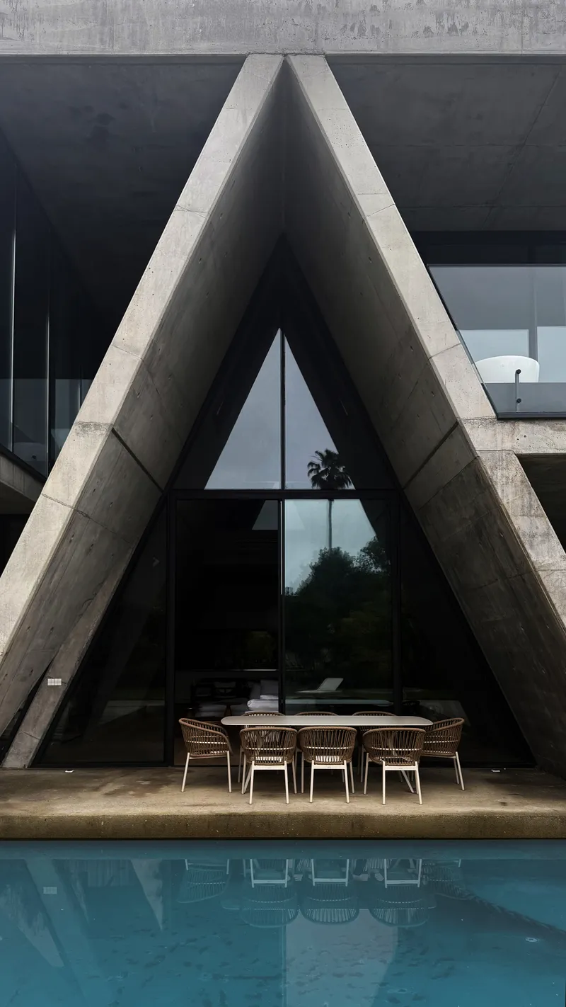 View of the triangular concrete portal framing the glazed entrance with a dining table beside the pool