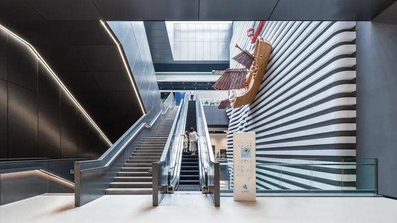 Interior atrium with escalator and staircase flanked by black paneled wall and horizontal striped wall with skylights above