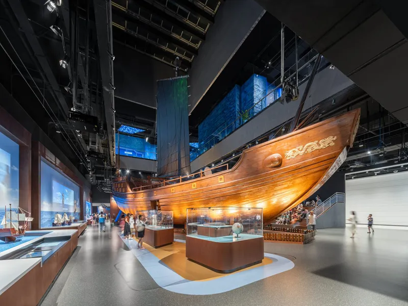 Museum gallery with suspended wooden ship hull above display cases and visitors on polished floor