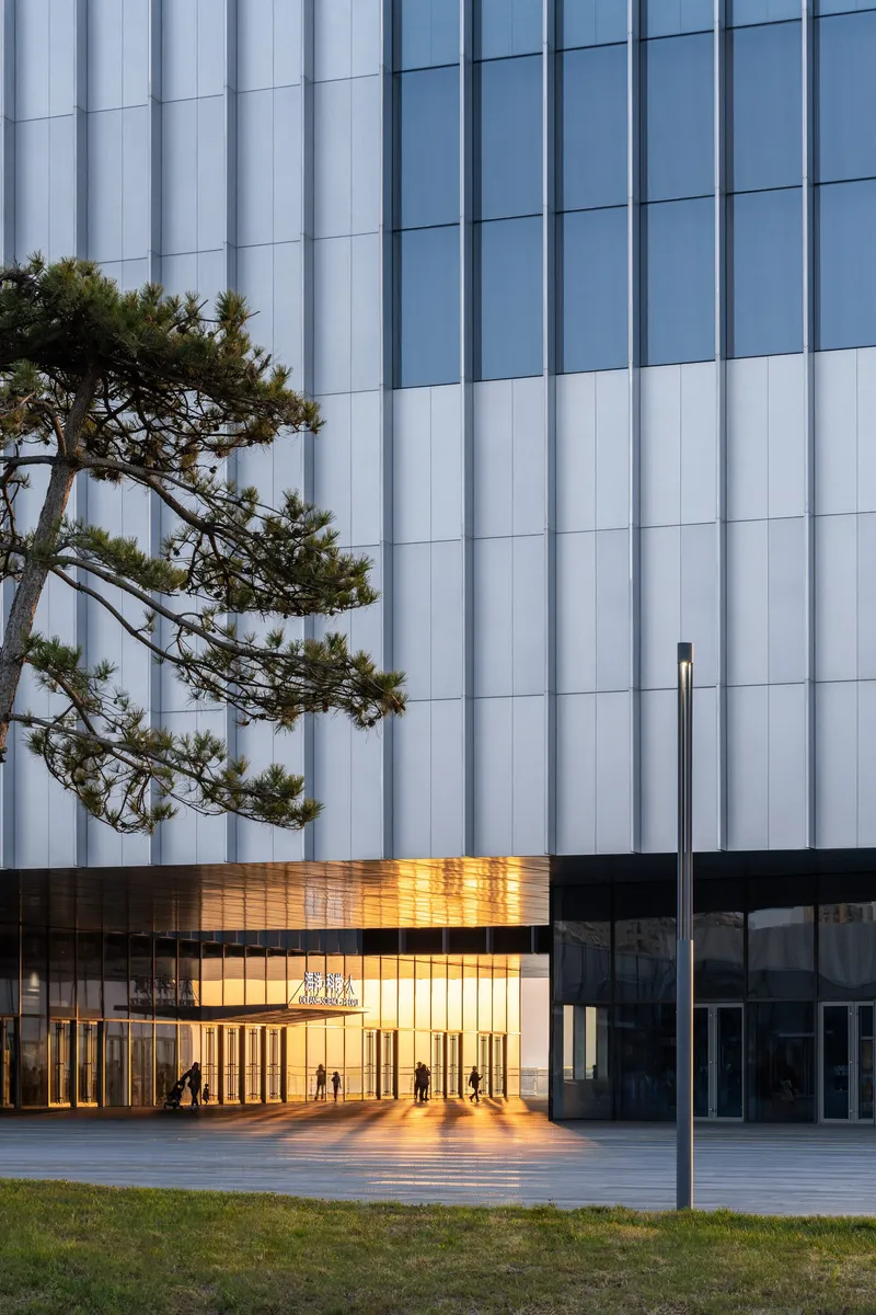 Illuminated glass entrance pavilion with vertical bronze fins at dusk beside pine tree and white tower facade