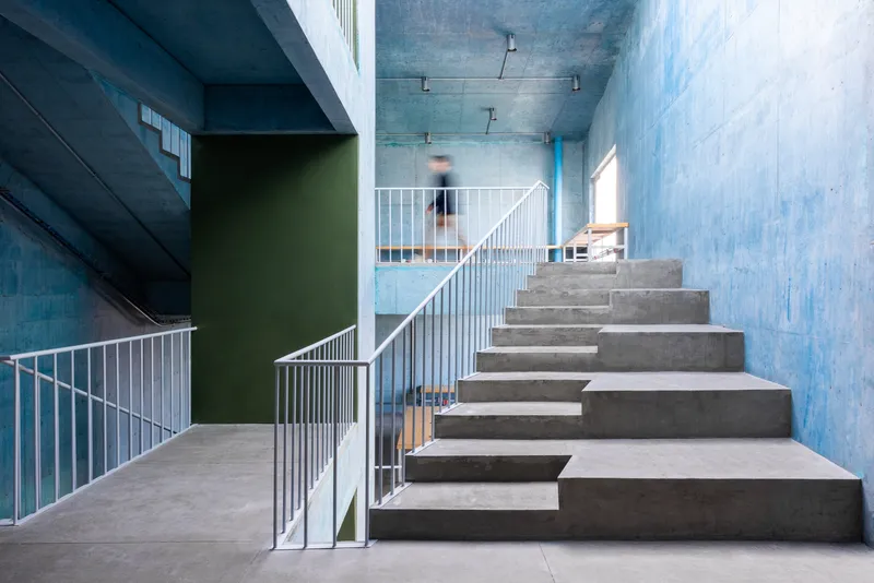Interior concrete staircase with white metal railings leading up through a triple-height space with blue-tinted walls