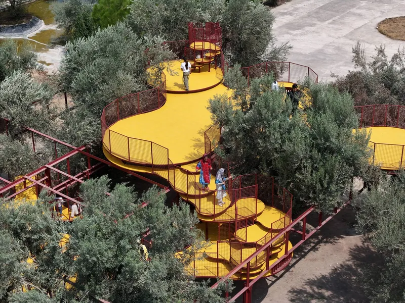 Elevated walkway with yellow mesh flooring and red steel framing threading through olive trees