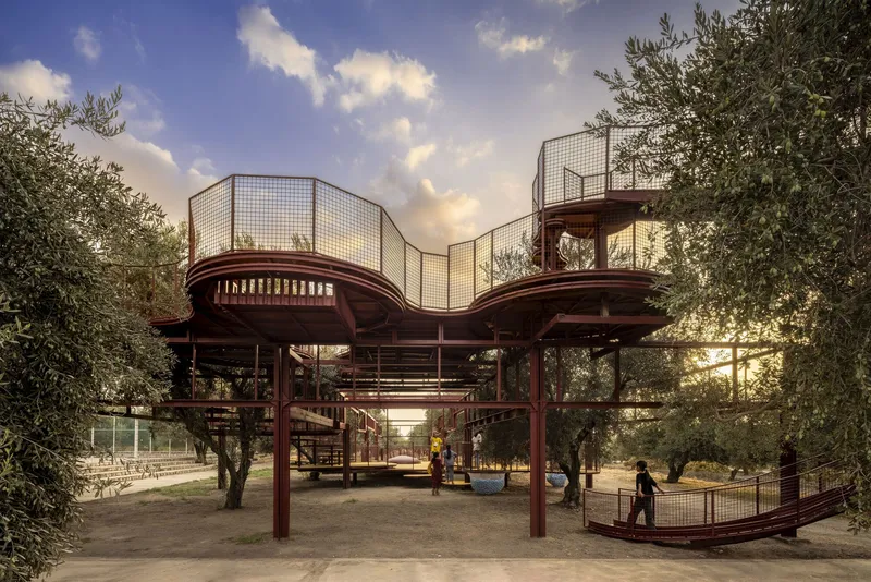 Elevated play structure with mesh screens and red steel framework at sunset among olive trees