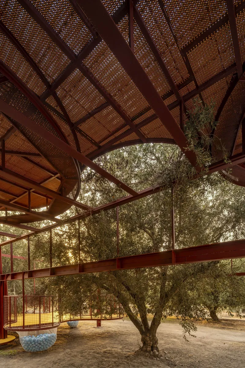 View upward through perforated metal panels and red steel beams framing an olive tree canopy