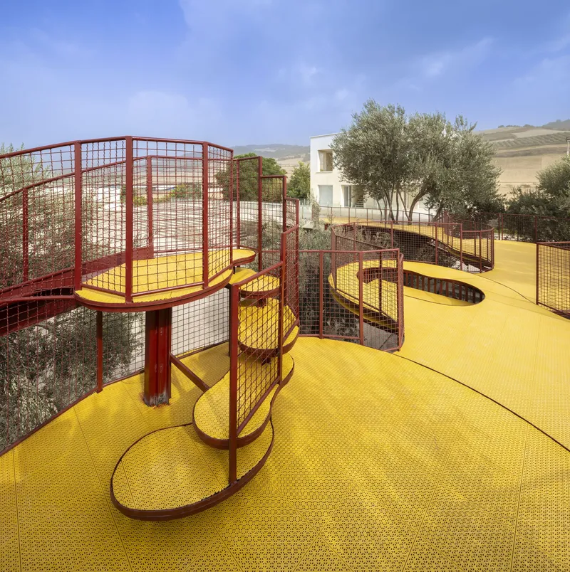 Elevated yellow walkway with red mesh railings curving past olive trees under clear skies
