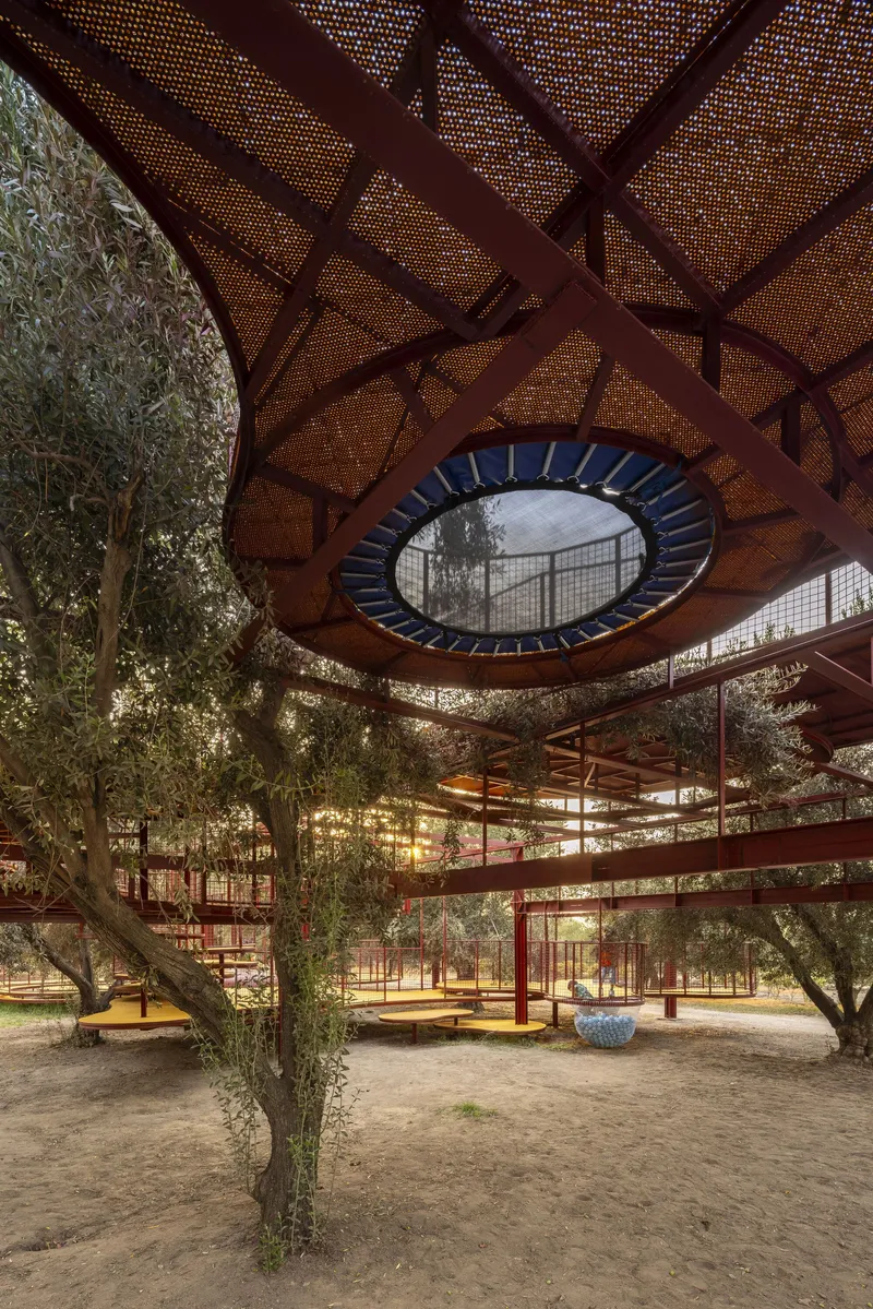 Underside of perforated metal canopy with circular opening and olive trees at dusk