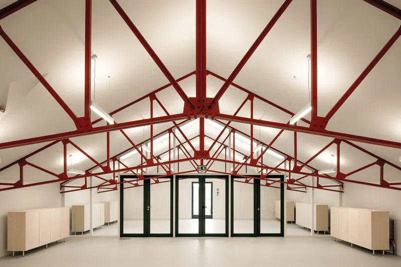 Interior hall with red steel truss system radiating from central ridge under vaulted white ceiling