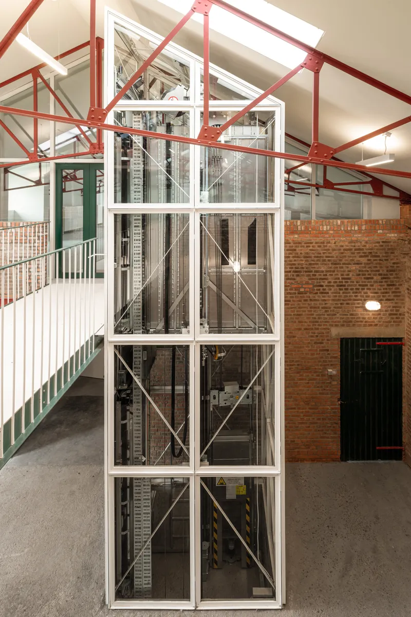 Glass-enclosed elevator shaft with exposed steel cross-bracing inside a warehouse with red painted trusses