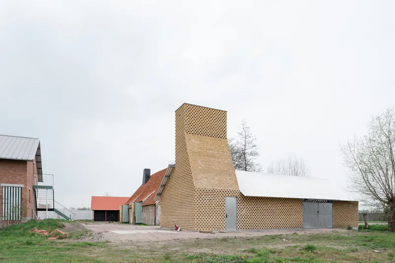 Perforated brick addition with tower chimney beside existing farm buildings under an overcast sky