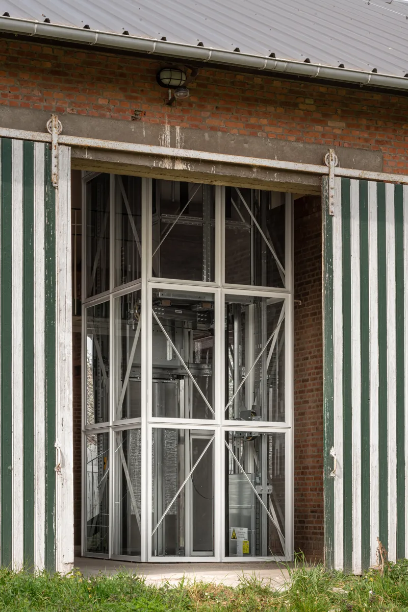 Exterior view of the glass elevator enclosure between green-and-white striped corrugated metal walls