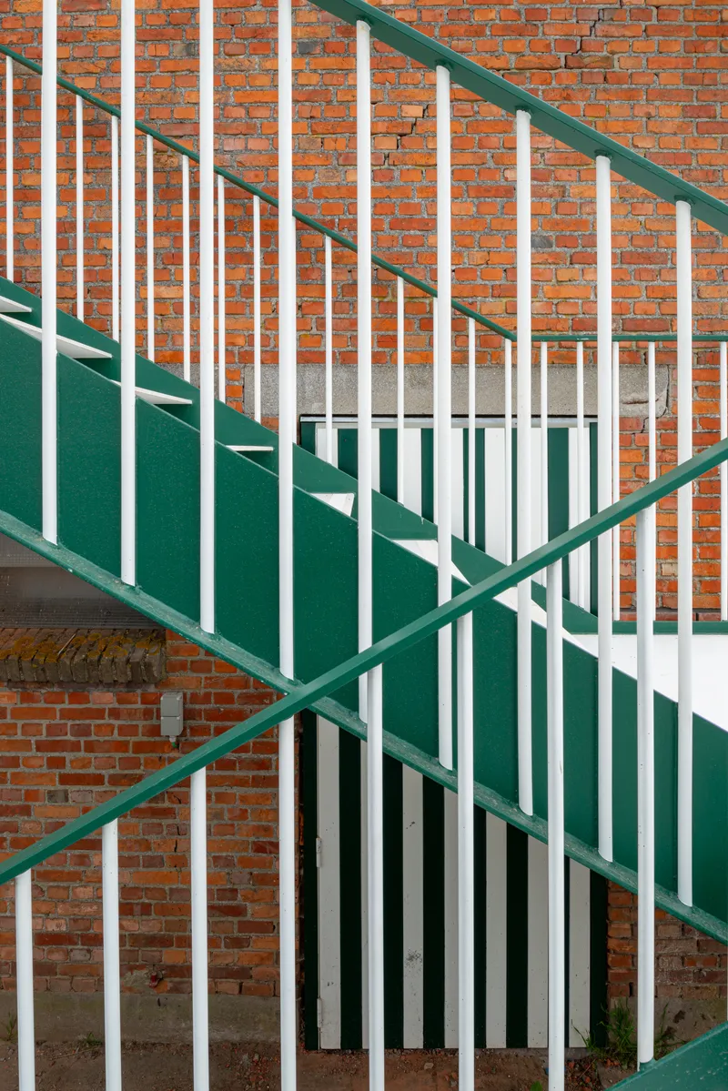 Green-and-white painted steel staircase with vertical bar railings ascending against exposed brick