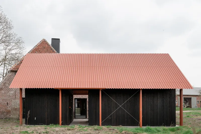 Black timber-clad barn volume with coral corrugated metal roof and orange steel columns under overcast sky