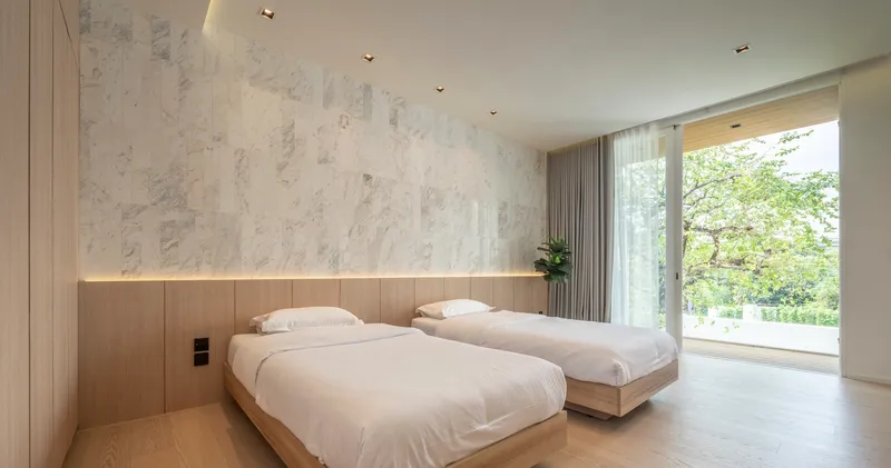 Bedroom with two timber-clad beds beneath a pale marble accent wall and garden views